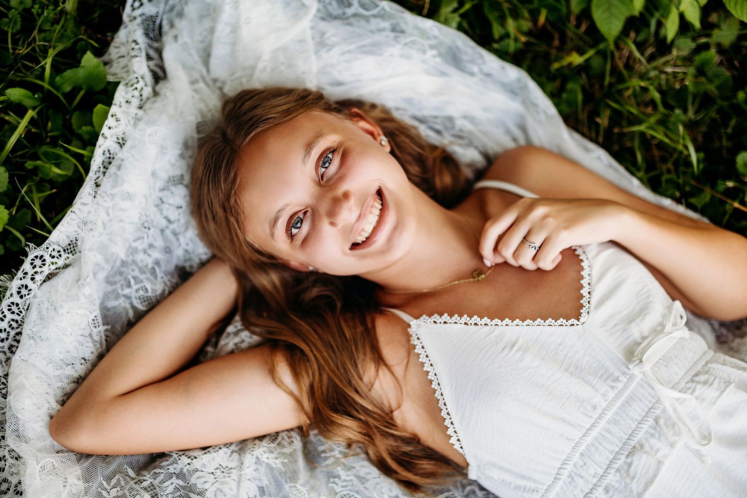 A person lies on white lace fabric in grass, smiling joyfully at the camera.