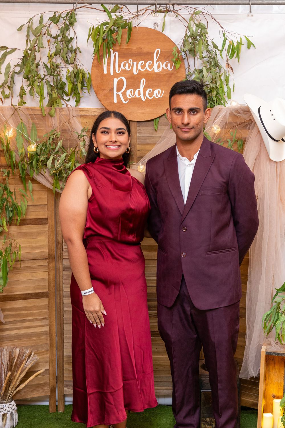 A couple in complementary burgundy and dark purple formal attire pose at an event with decorative greenery backdrop.