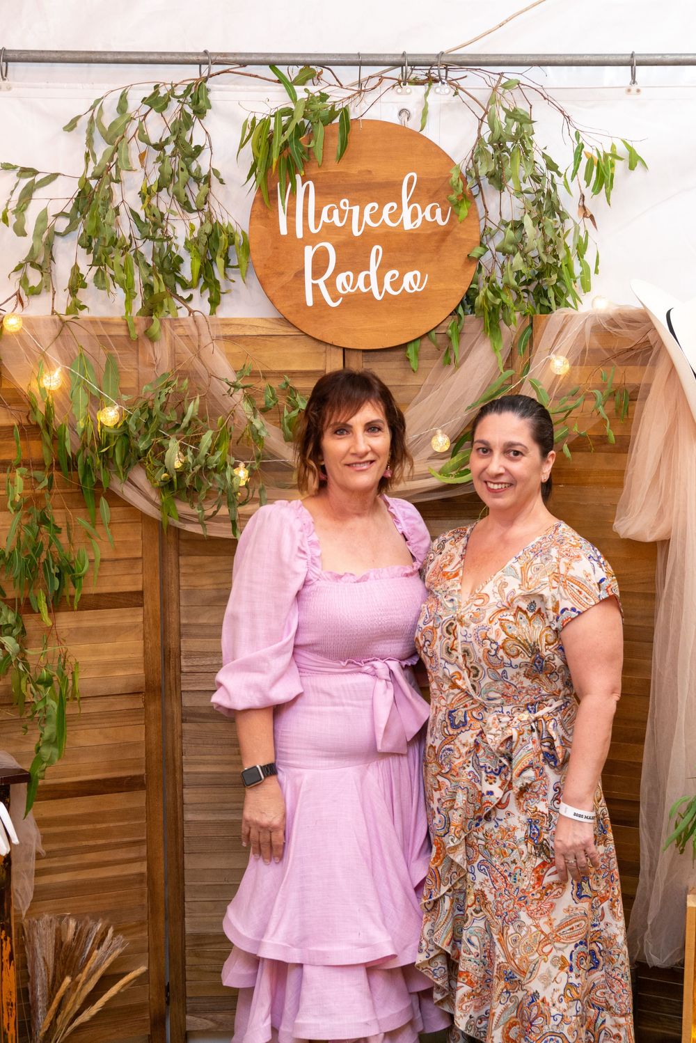 Two women in pink and floral dresses pose together at an outdoor event with wooden wall and greenery decorations.