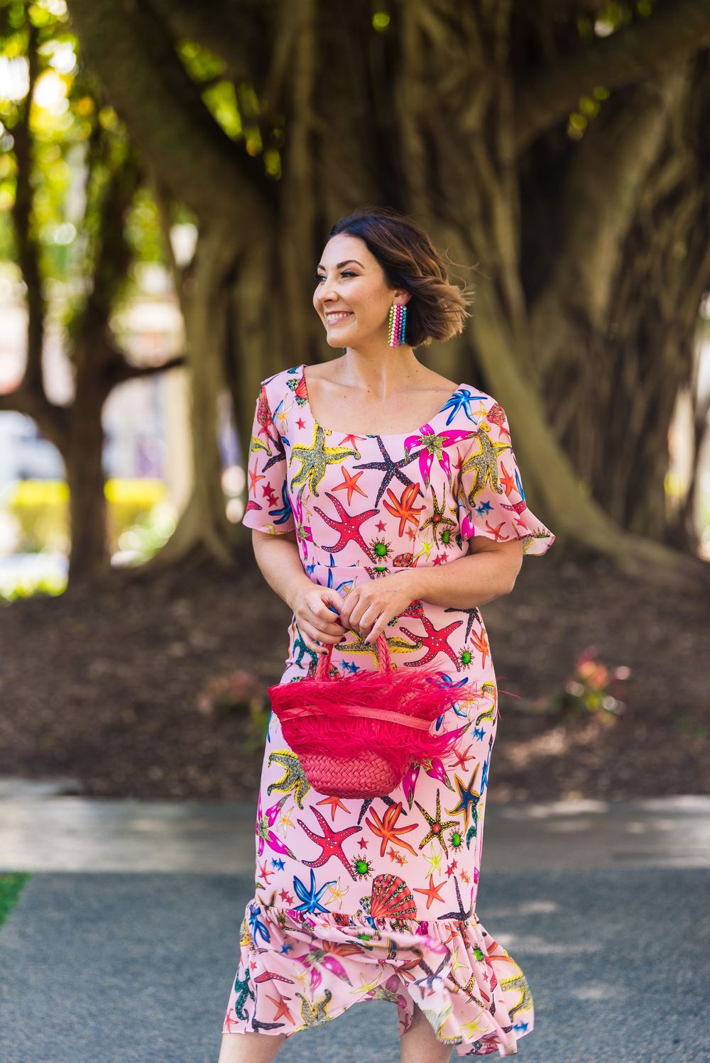 Woman in vibrant floral print dress with pink clutch walking along a tree-lined path on sunny day.