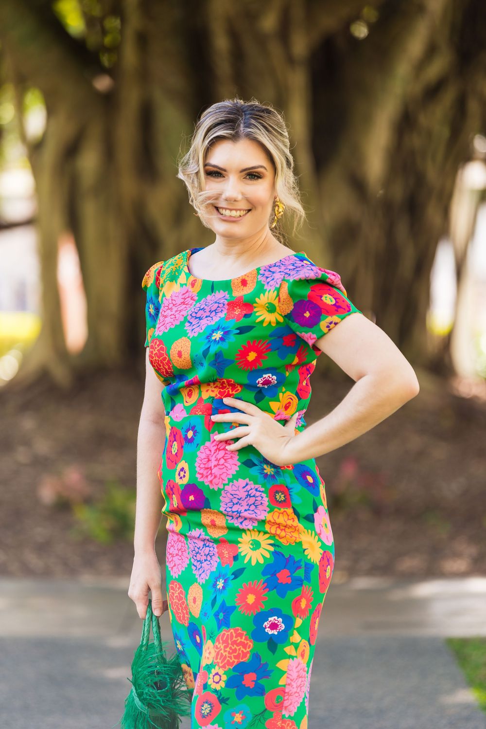 Woman in vibrant floral dress smiles and poses outdoors against a blurred tree background.