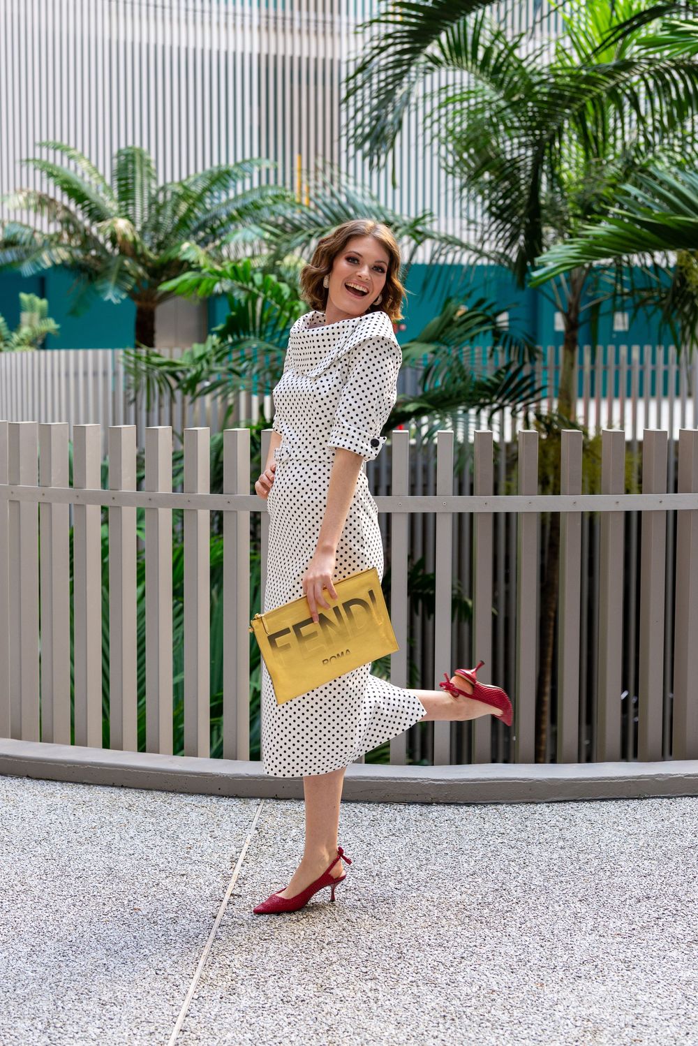 Woman in polka dot dress twirls playfully with a yellow handbag against a grey and tropical backdrop.