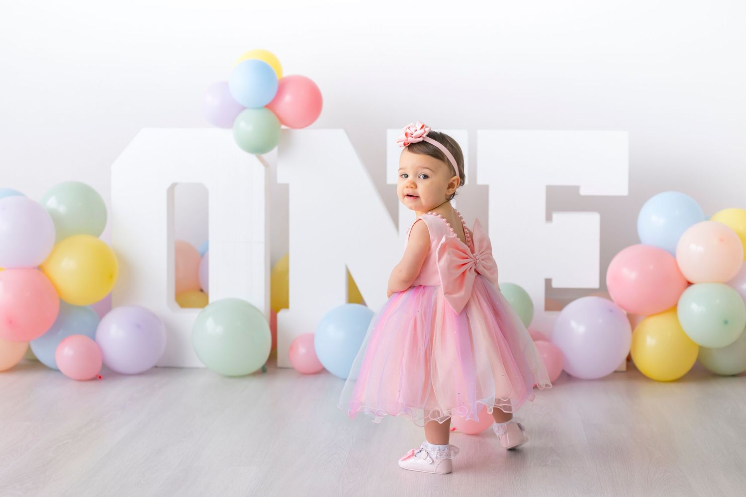 A small child in a pink tulle dress stands by decorative ONE letters with pastel balloons on a white backdrop.