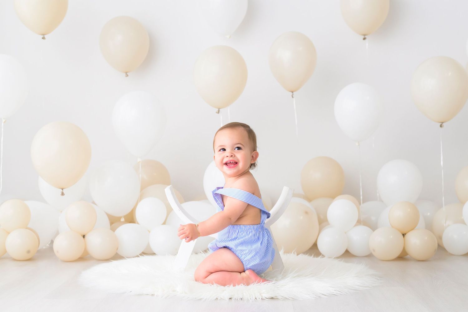 A baby in a blue dress sits on a white floor surrounded by cream and white balloons for a photo session.