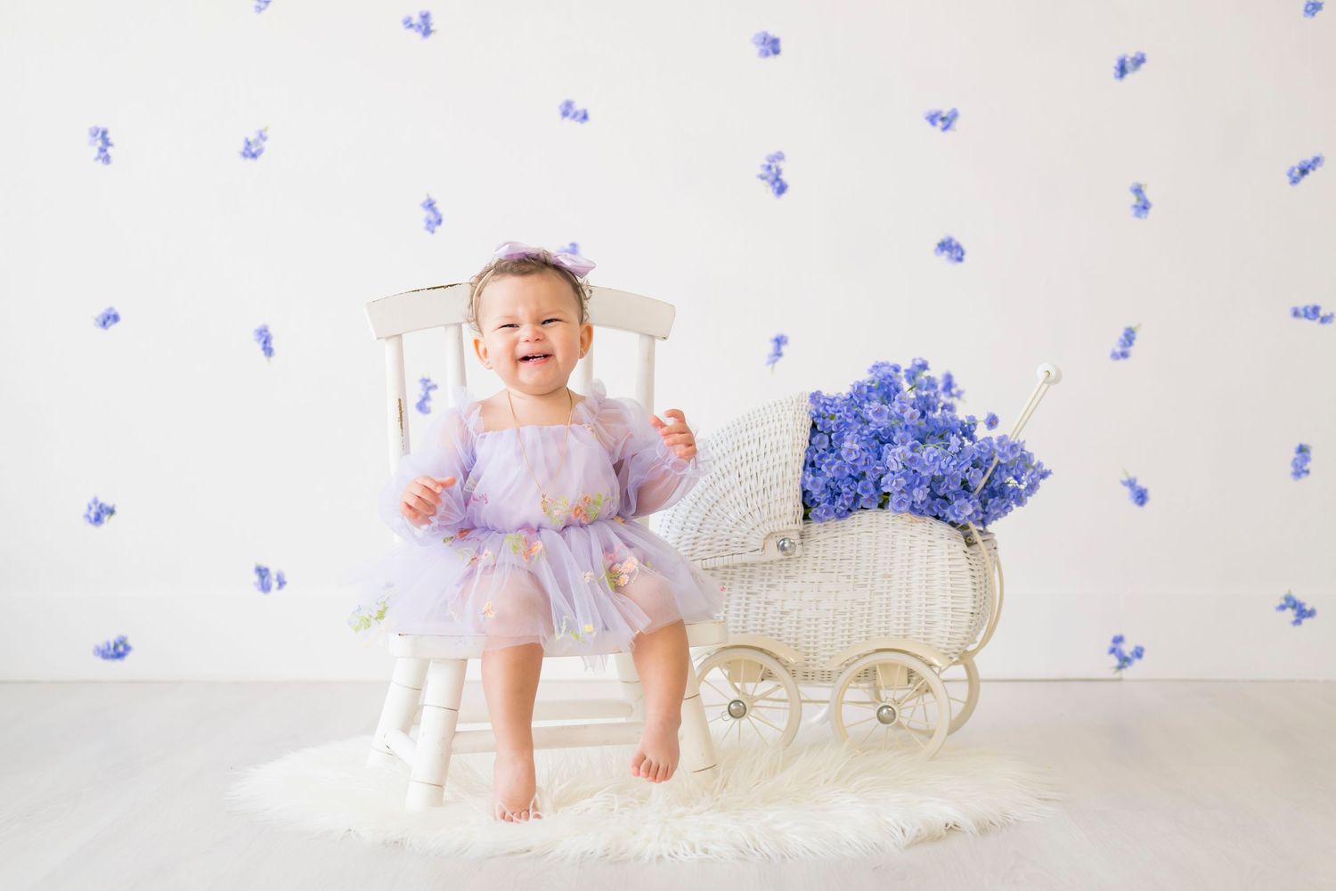 Toddler in a tulle dress sits on white chair with blue floral decor and floral arrangement in elegant setup.