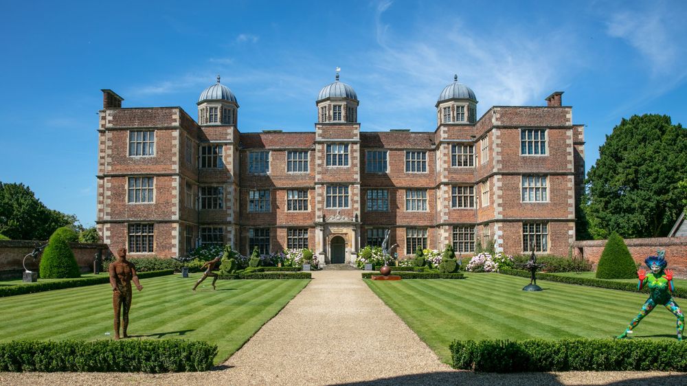 The front of the red brick Elizabethan frontage of Doddington Hall bathed in sunshine with blue skies, sculptures adorn the lawns