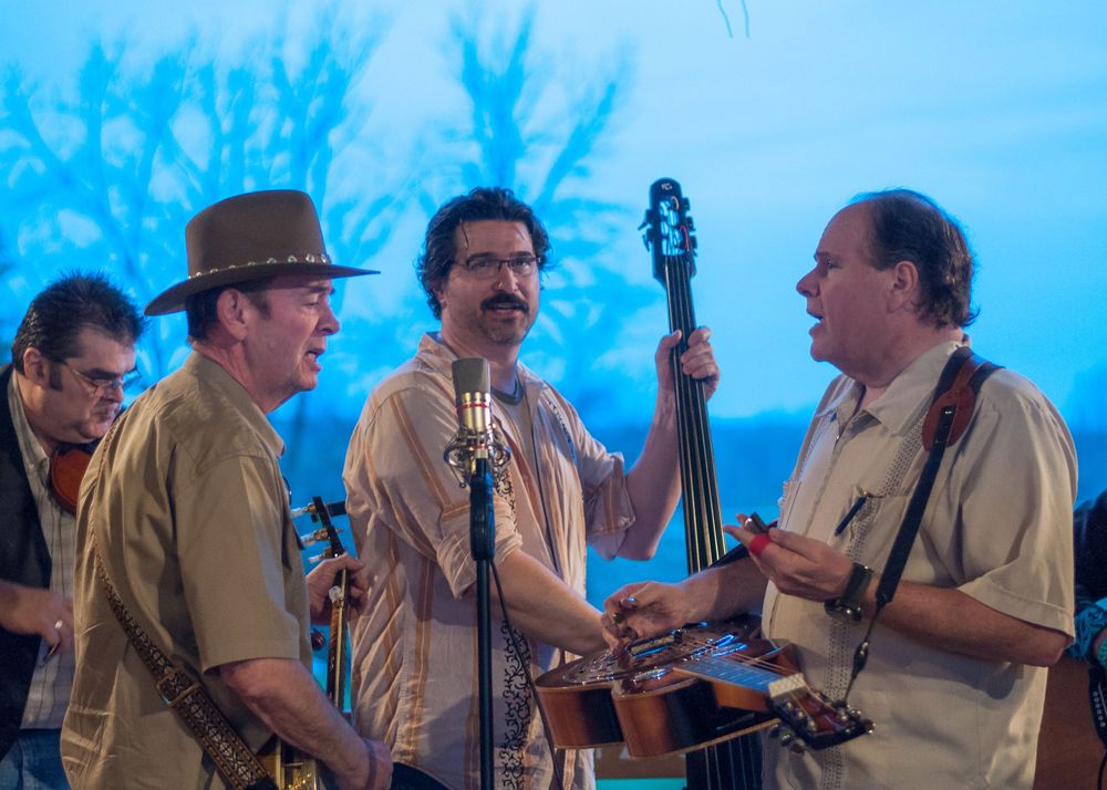 Musicians performing bluegrass music outdoors against a bright blue sky backdrop.