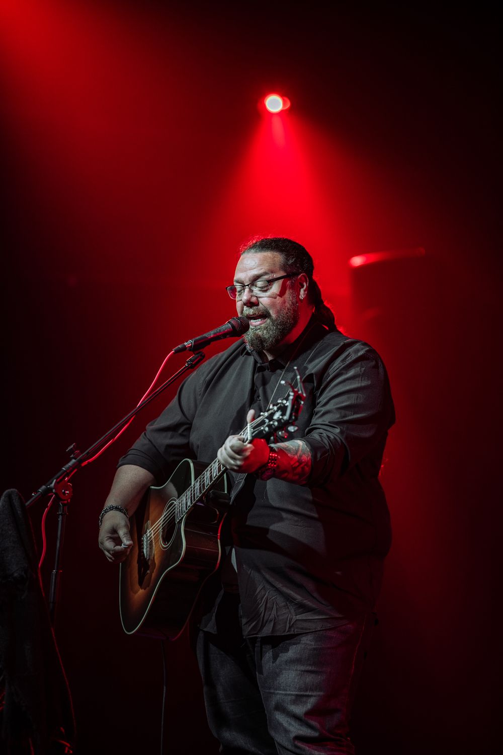 Concert performer plays acoustic guitar under dramatic red stage lighting during live performance.
