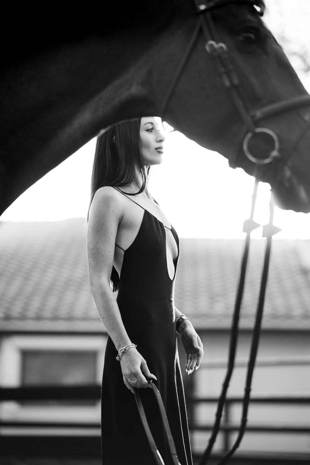 Elegant woman in black dress stands beside a dark horse holding reins in a black and white equestrian portrait.