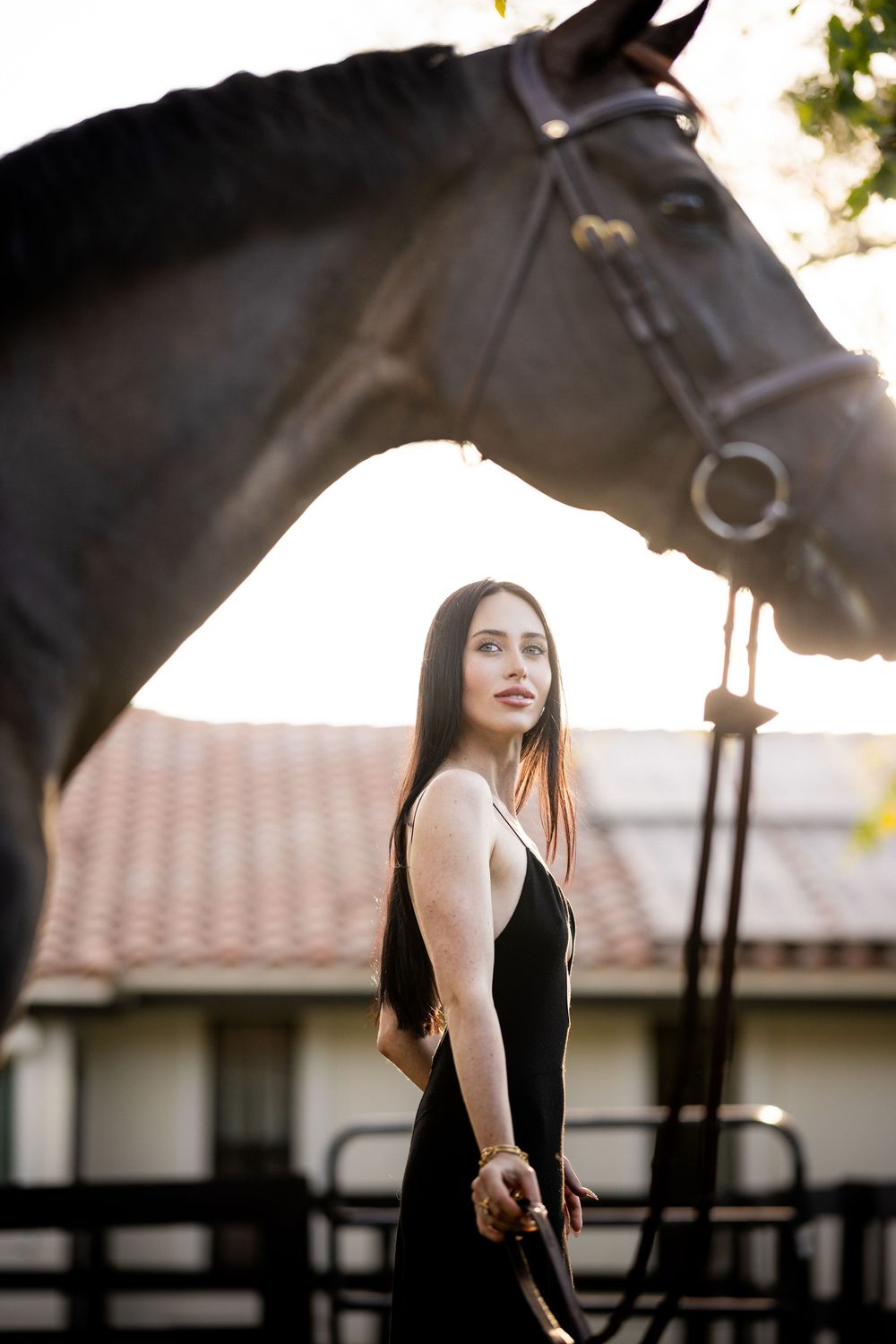 Young woman in black dress poses with dark horse at golden hour near stable.