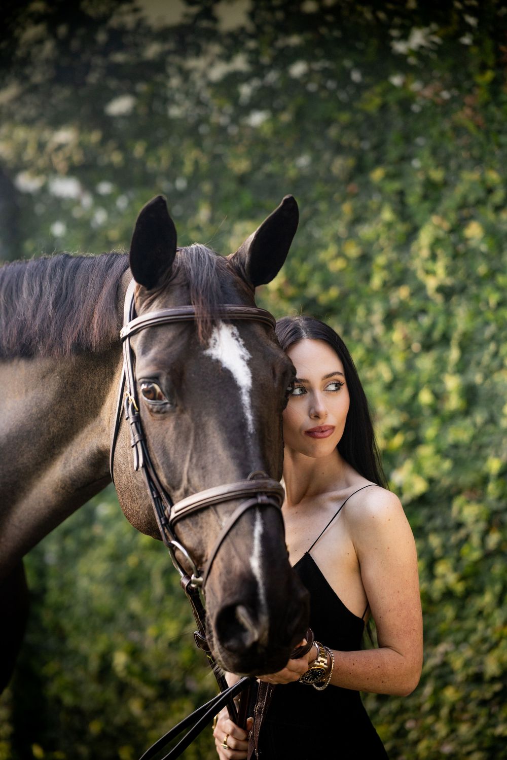 Beautiful brunette woman in black dress poses closely with a dark horse wearing a bridle outdoors.
