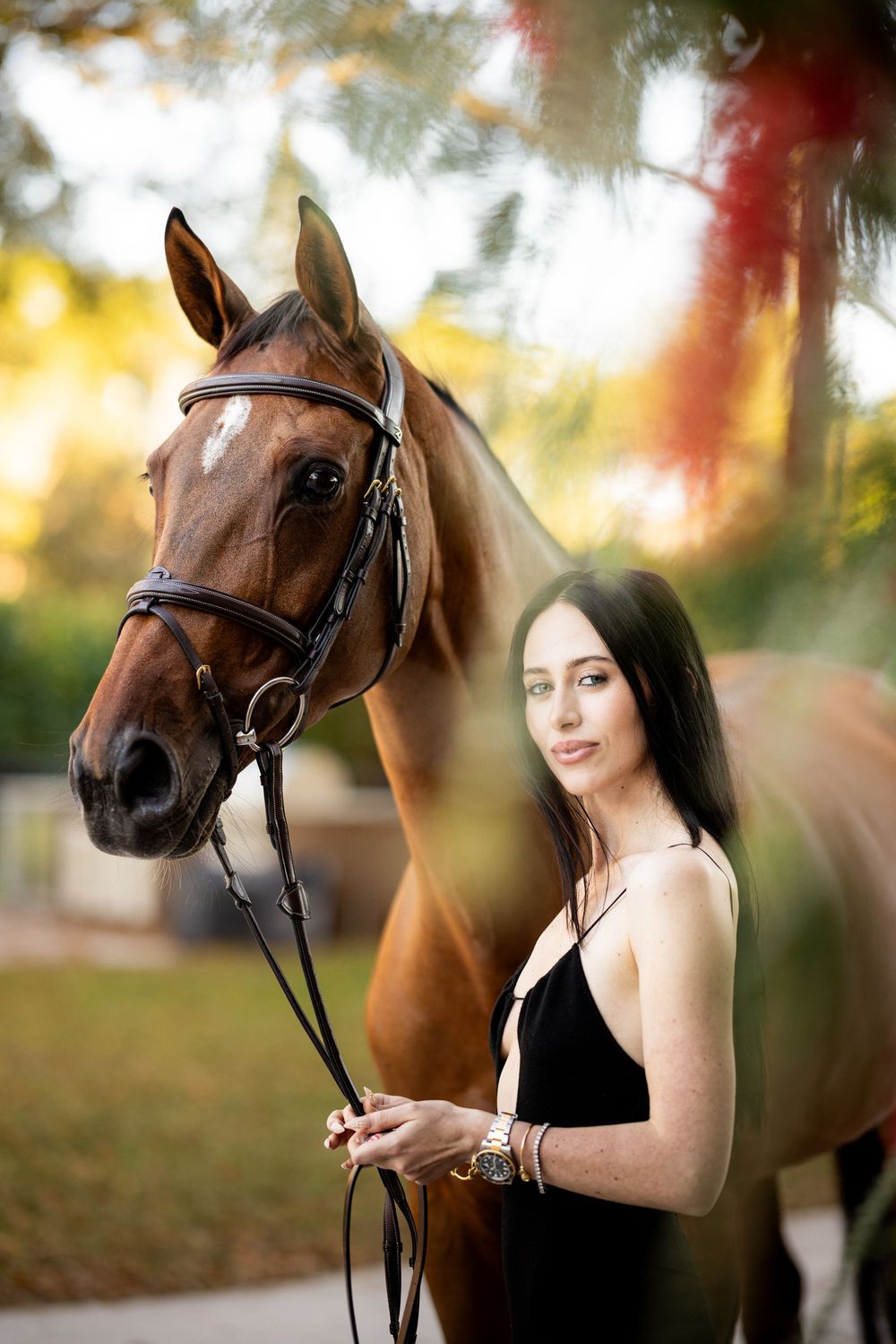 Young woman in black dress stands beside a chestnut horse with bridle outdoors at golden hour.