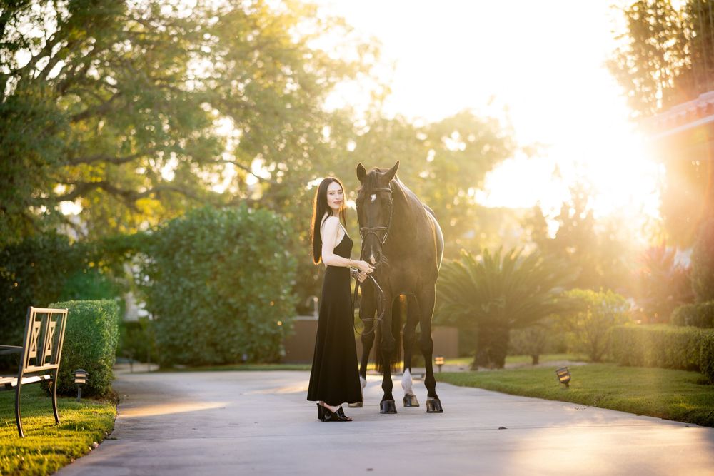 Woman in elegant black dress walks with dark horse along sunlit garden path.