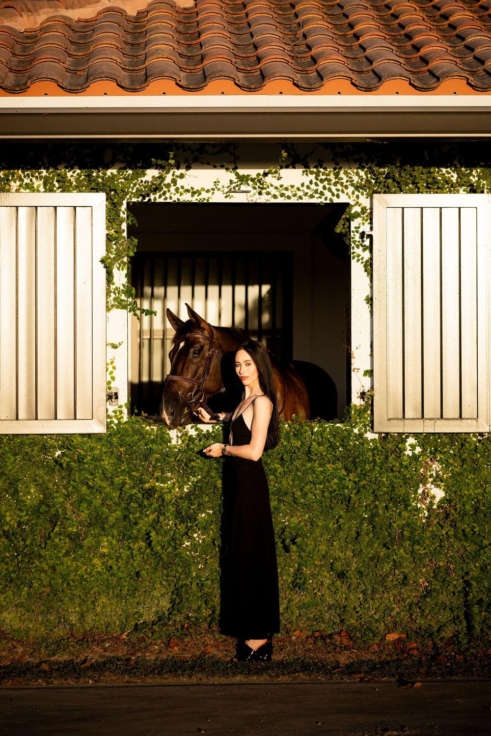 Woman in black gown standing beside a dark horse peering from an ivy-covered stable window.