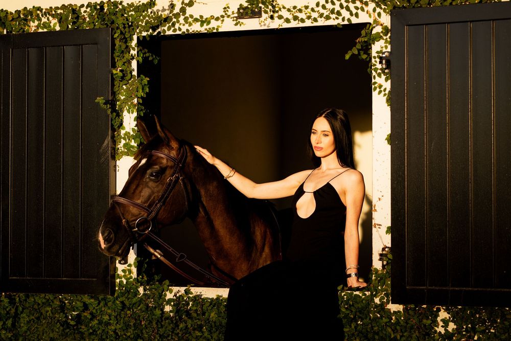 Brunette woman in black dress gently touching a dark horse emerging from stable doors.