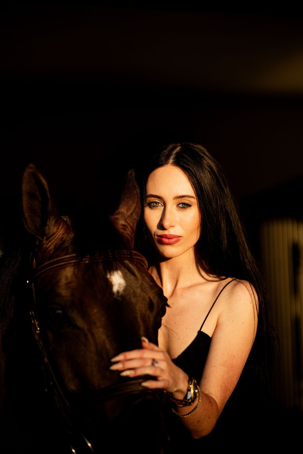 Portrait of a dark-haired woman in black dress tenderly holding a horse in dramatic low light.