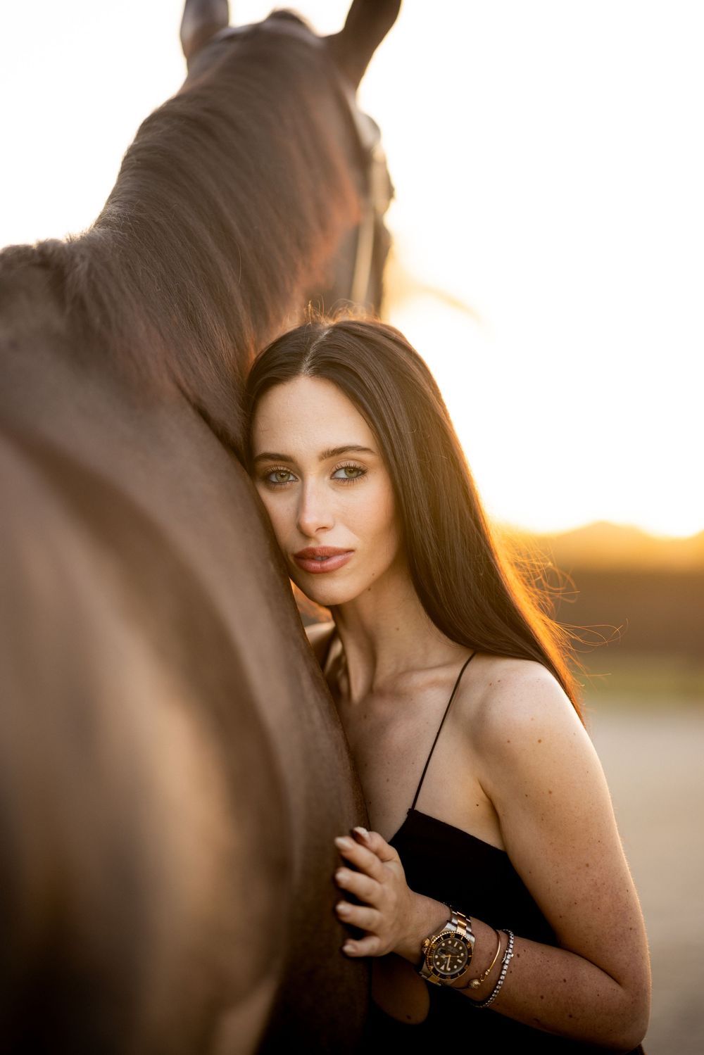 A woman in a black dress embraces a dark horse at golden hour, her long hair glowing in warm sunset light.