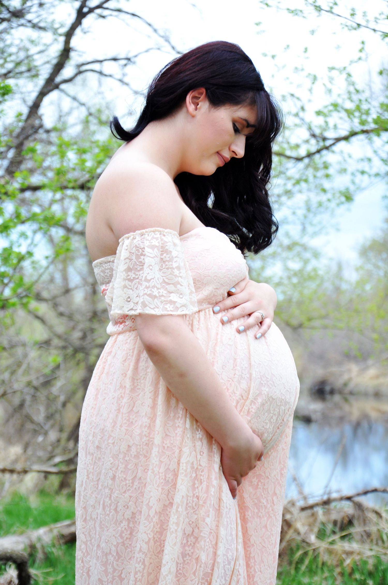 Maternity photo session in a pale pink lace dress by a lake during spring.