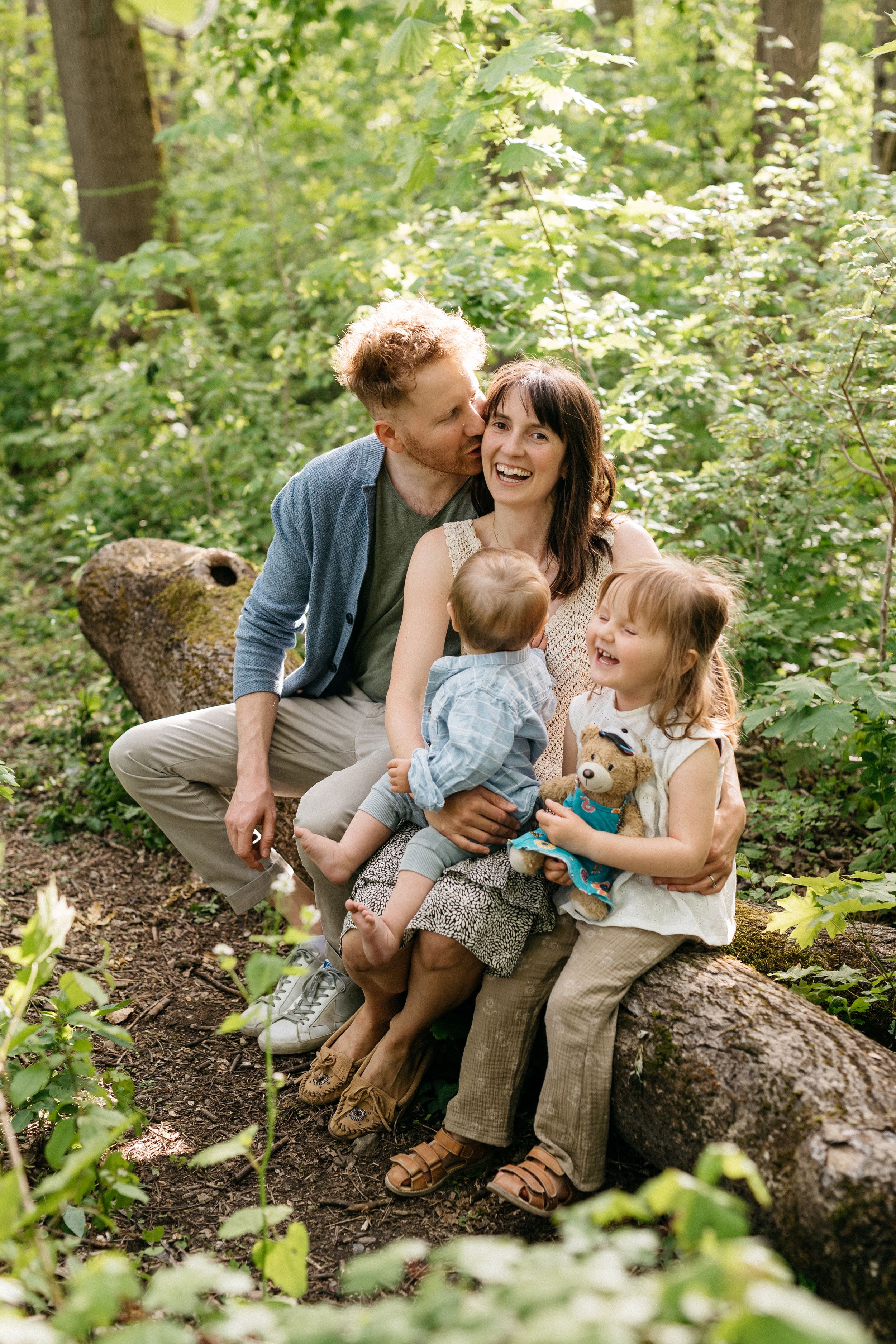 Natürliches ungestelltes Familienfoto draußen im Park. Mama, Papa und zwei Kinder kuscheln auf Baumstamm.