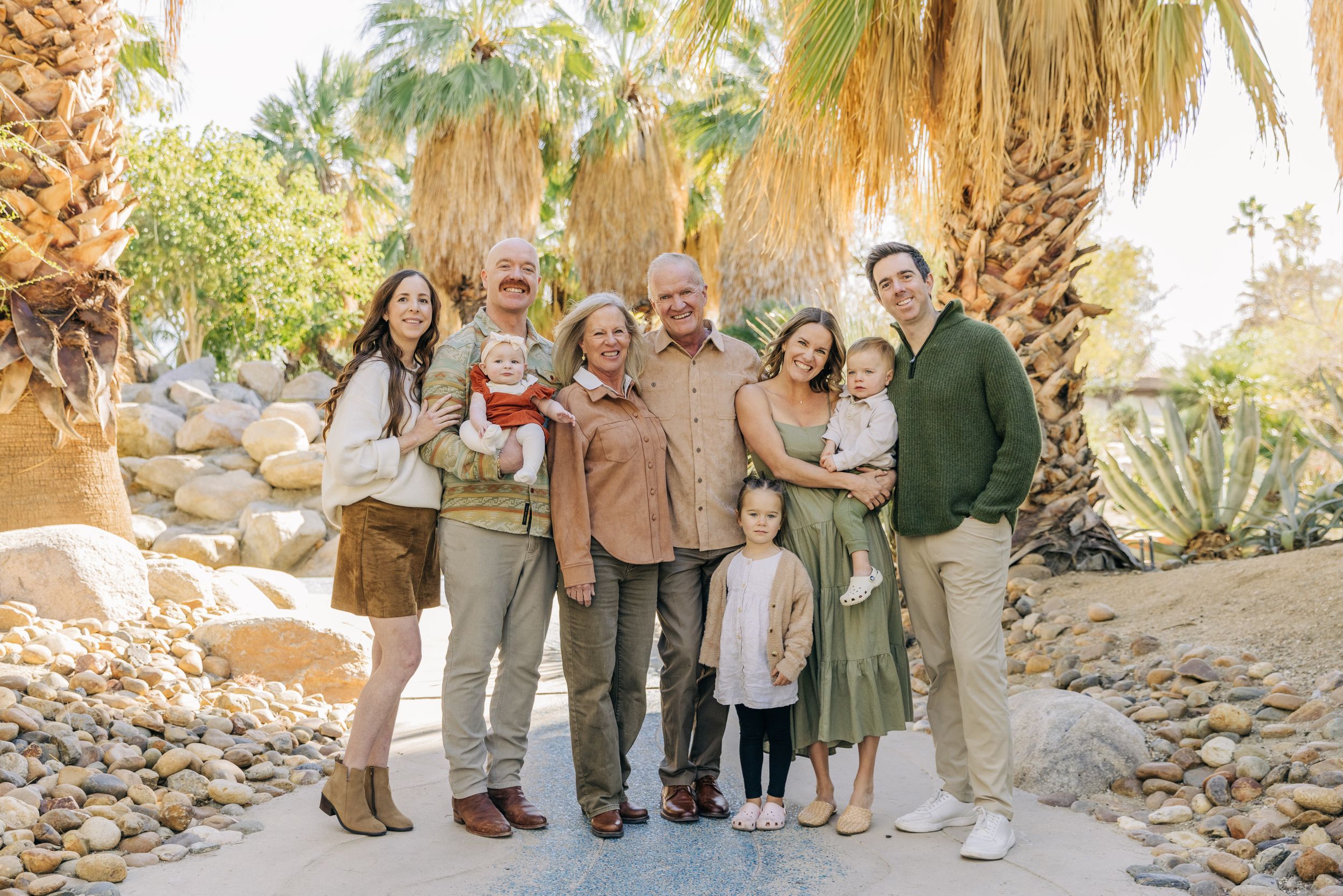 A group poses for family photos in a desert landscape with palm trees and rocky terrain at sunset.