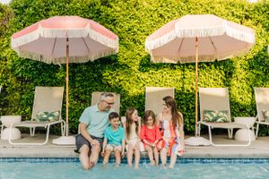 Family relaxes together on poolside lounge chairs under pink umbrellas next to a lush green hedge wall in Palm Springs