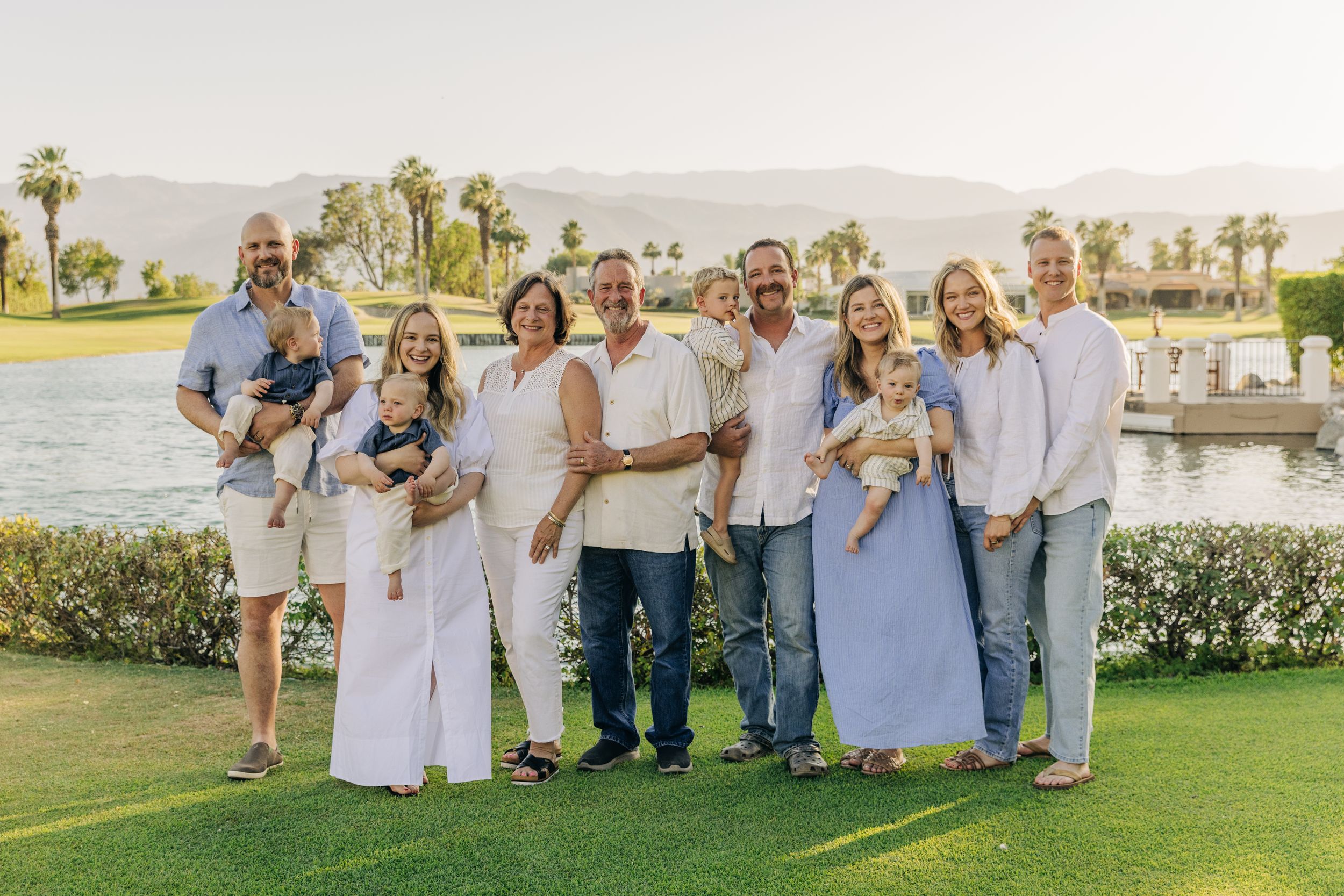Multi-generational family portrait outdoors in Palm Desert near a lake with palm trees and mountains in the background.