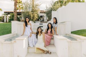 Four friends in casual summer outfits pose together on white stone steps in a sunny garden setting.