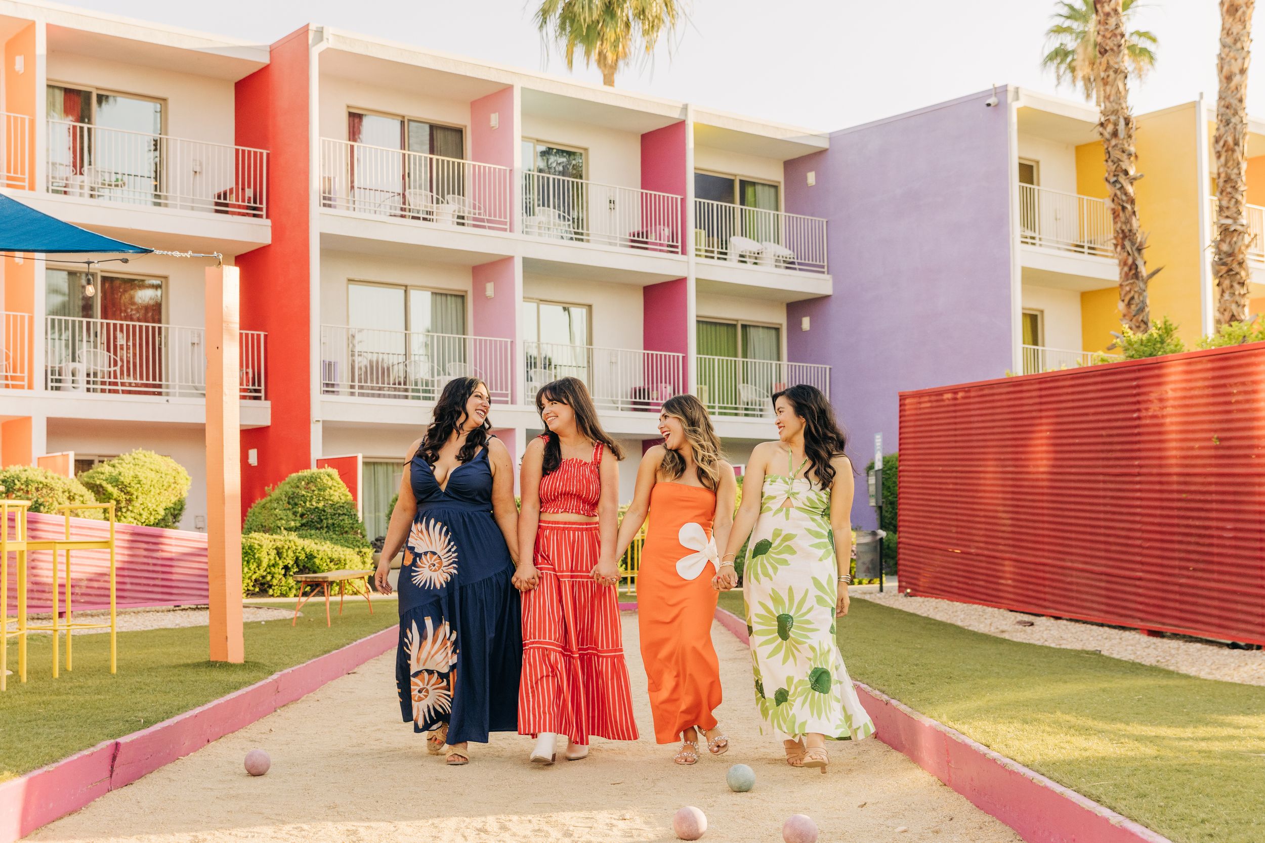 Four women in colorful maxi dresses walk together outside Saguaro Hotel in Palm Springs, a vibrant, multi-colored hotel with pink accents.