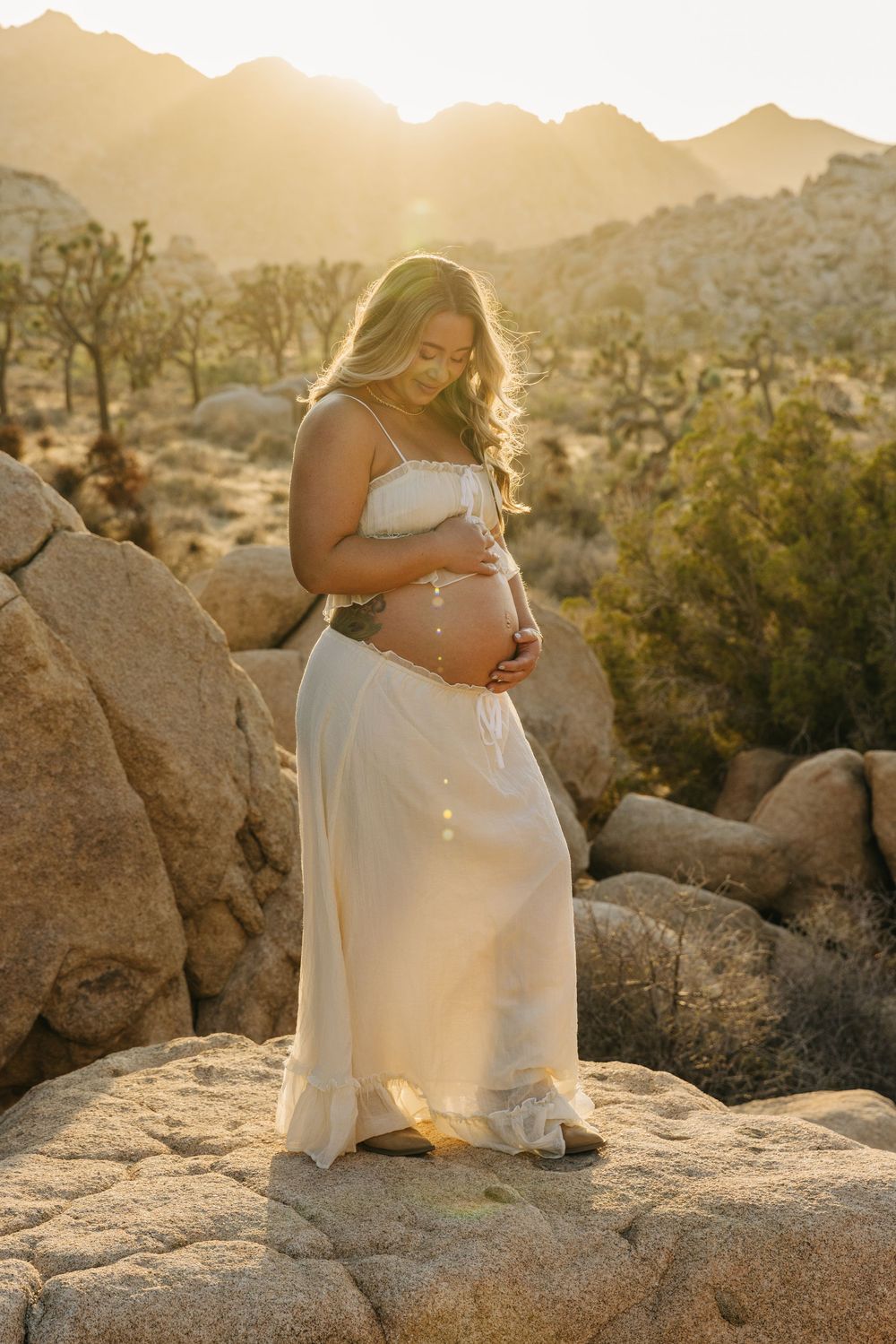 Maternity photoshoot at sunset in Joshua Tree National Park with a woman in white dress sitting on desert rocks.