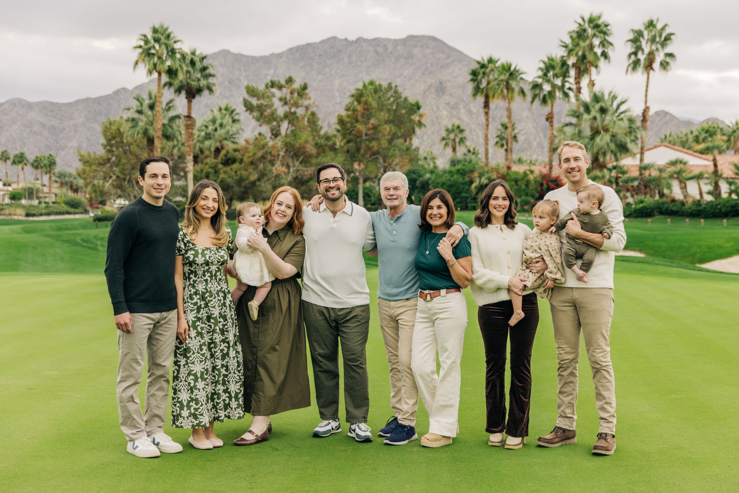 A group of people pose together on a sunny golf course with palm trees and mountains in the background.