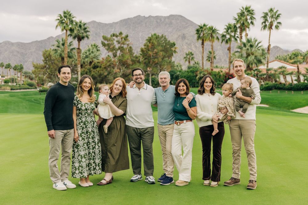 A group of people pose together on a sunny golf course with palm trees and mountains in the background.