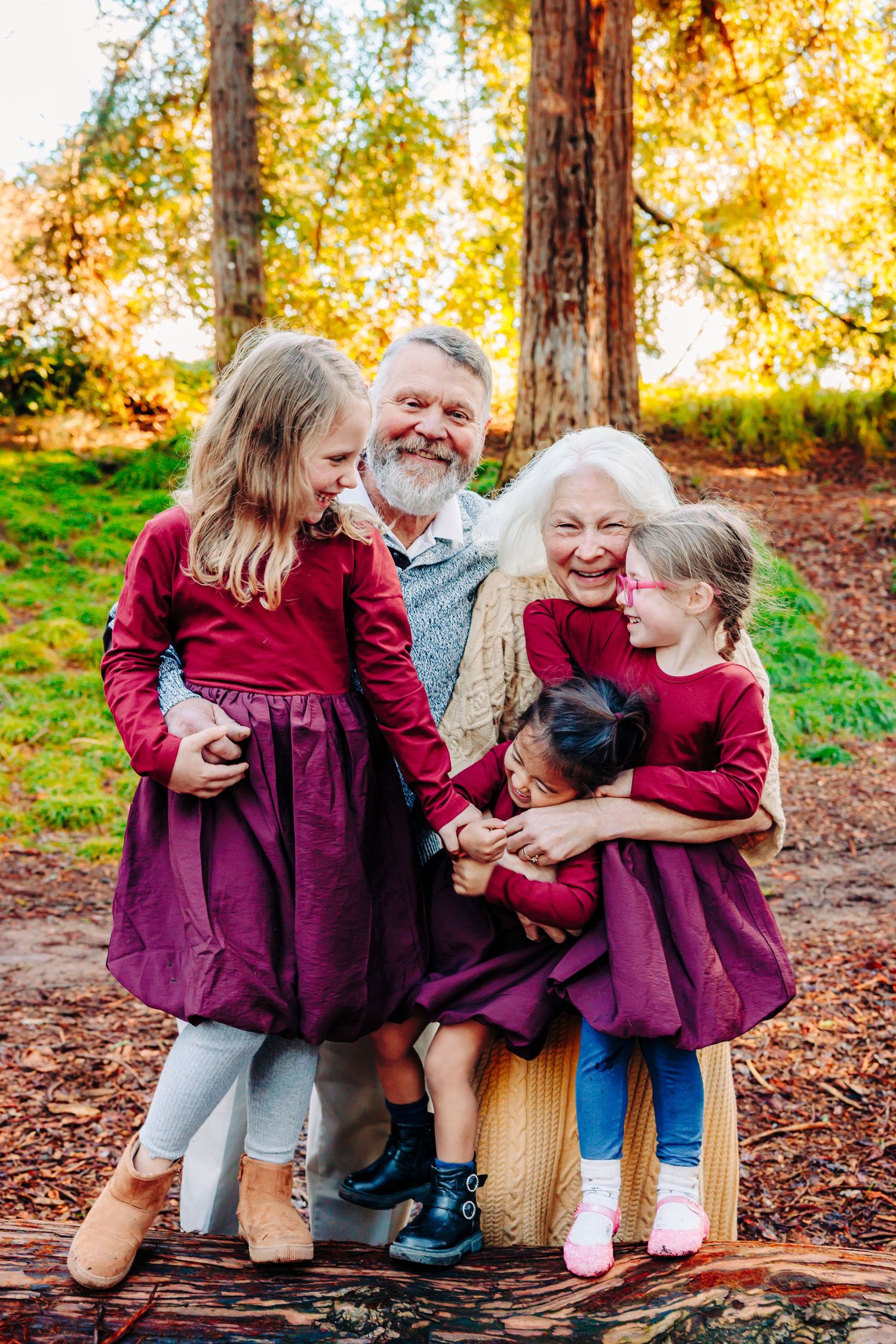 Family wearing burgundy outfits shares joyful laughter while sitting on wooden bench surrounded by autumn foliage.