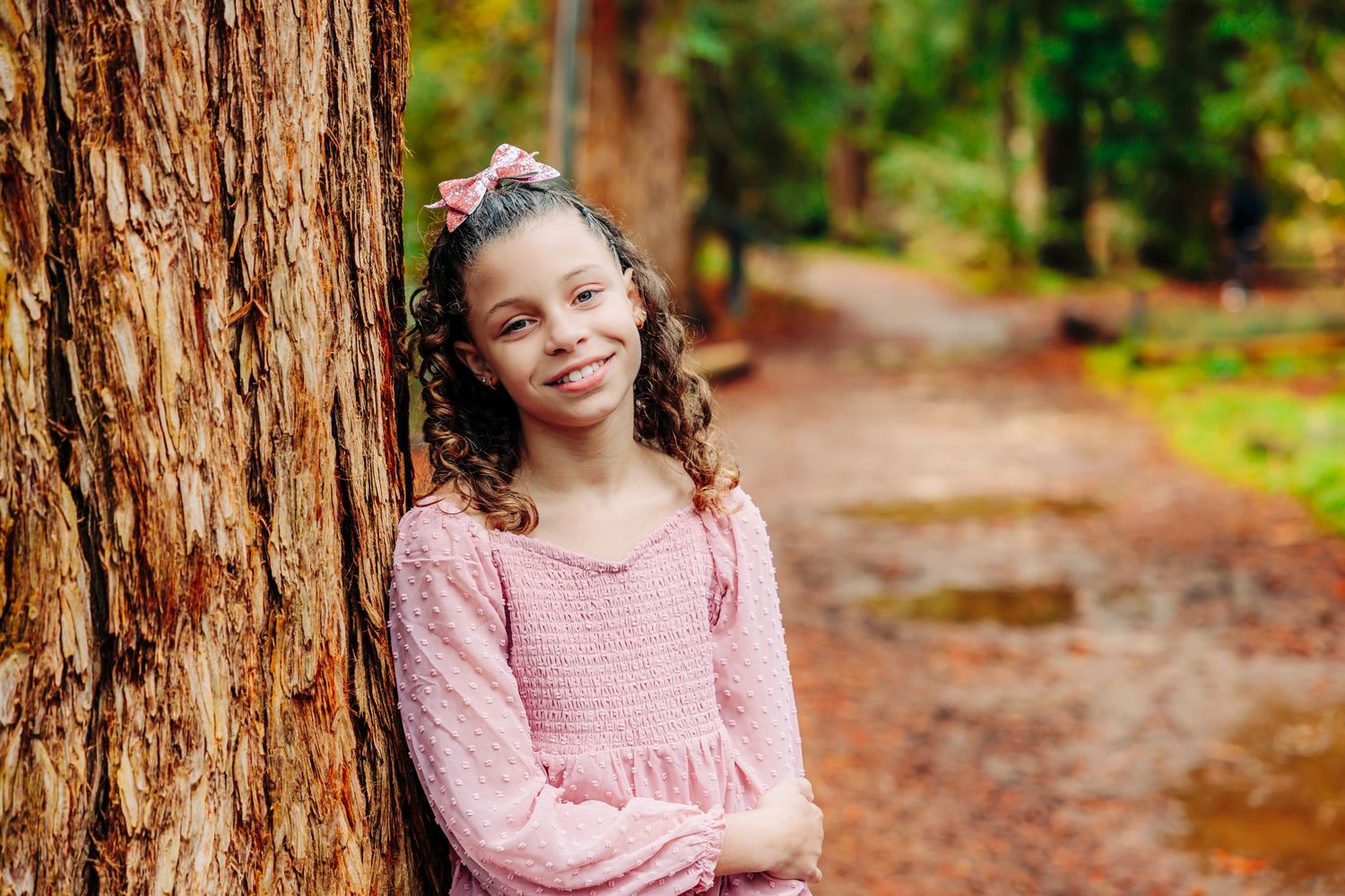 A young figure in a pink sweater poses against a tree trunk on a wooded autumn path with fallen leaves.
