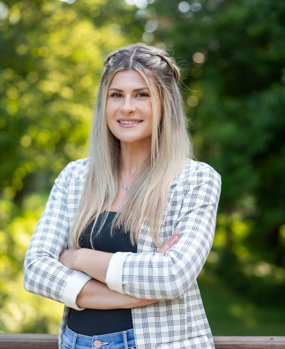 Woman in black top and plaid shirt poses outdoors with natural bokeh background.