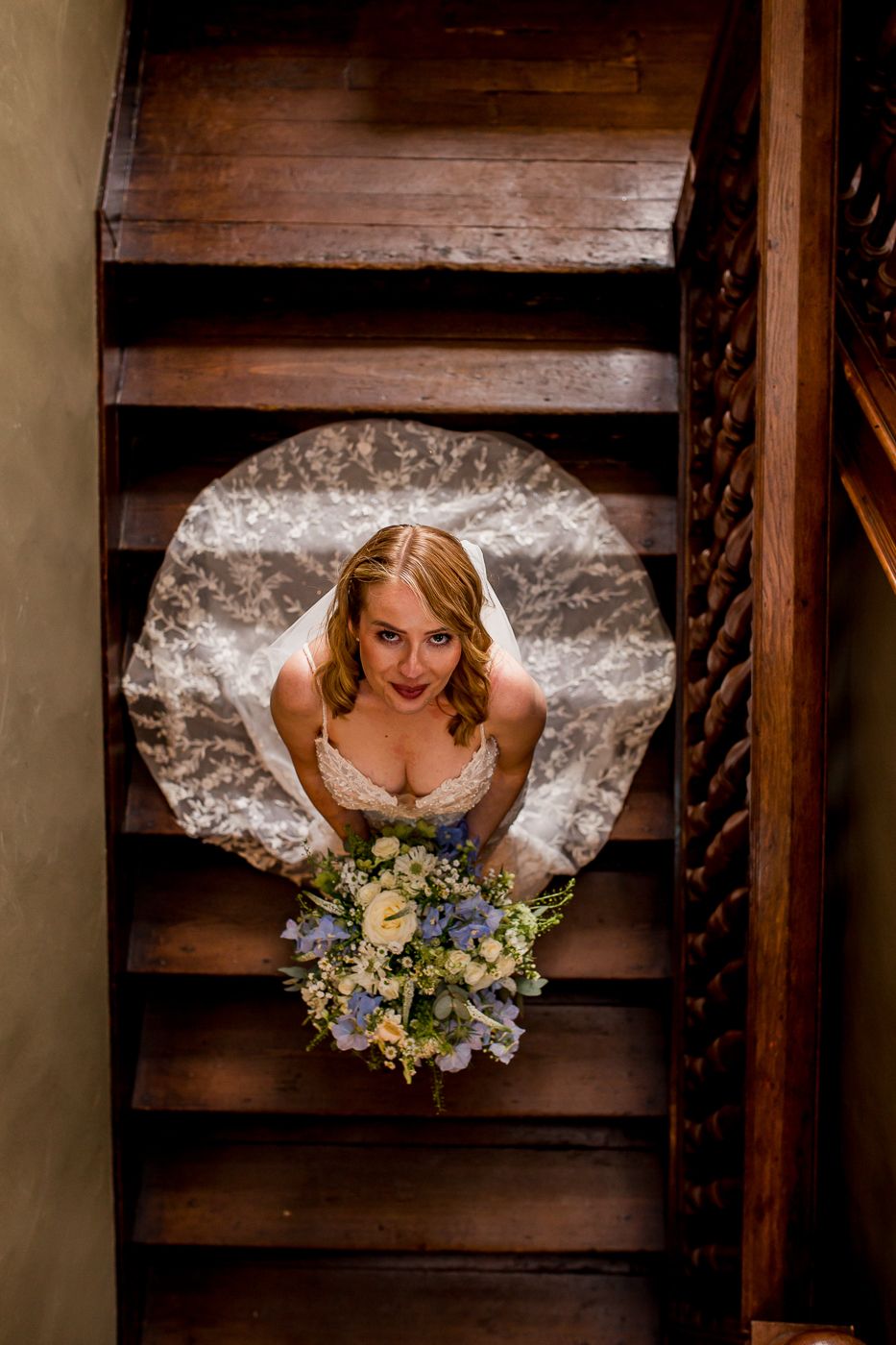 Artistic overhead view of bride in white dress with floral bouquet on wooden staircase.