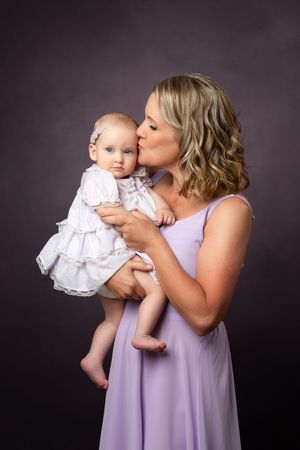 A woman in a lavender dress holding and kissing a baby wearing a white lace christening gown against a dark backdrop.