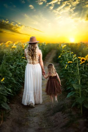 Two figures in flowing dresses walk hand in hand through a sunlit sunflower field at golden hour.