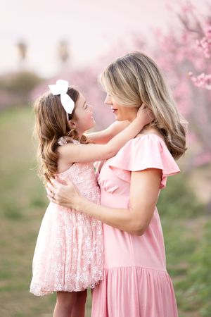 Two people wearing pink dresses embrace tenderly among blooming cherry blossom trees in a springtime garden setting.