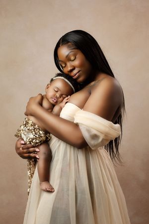 A woman in an ethereal cream dress holds her sleeping baby tenderly against a neutral backdrop in a studio portrait.