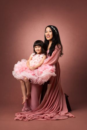 A mother and daughter pose together in matching pink dresses against a pink background in a professional studio portrait.