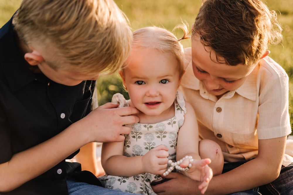 Two older boys lovingly cuddle a smiling baby girl outdoors in warm golden sunlight. childhood siblings photography in pleasant view tn