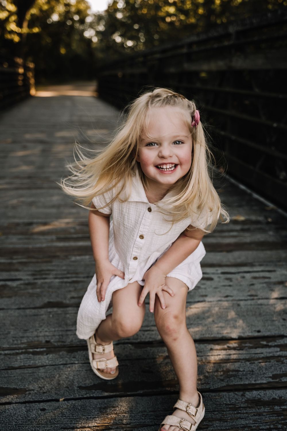 A joyful blonde toddler girl in a white dress crouches on a wooden bridge in nashville tn, smiling brightly with hair blowing in the wind.