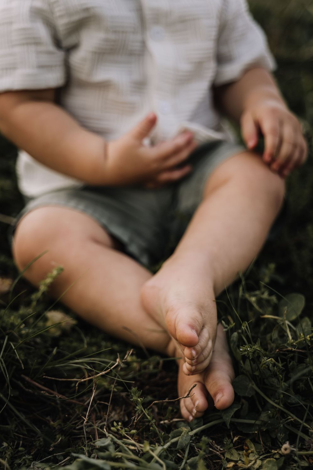 Barefoot toddler sitting cross-legged on green grass in white shirt and gray shorts. details photographduring a littles milestone photoshoot
