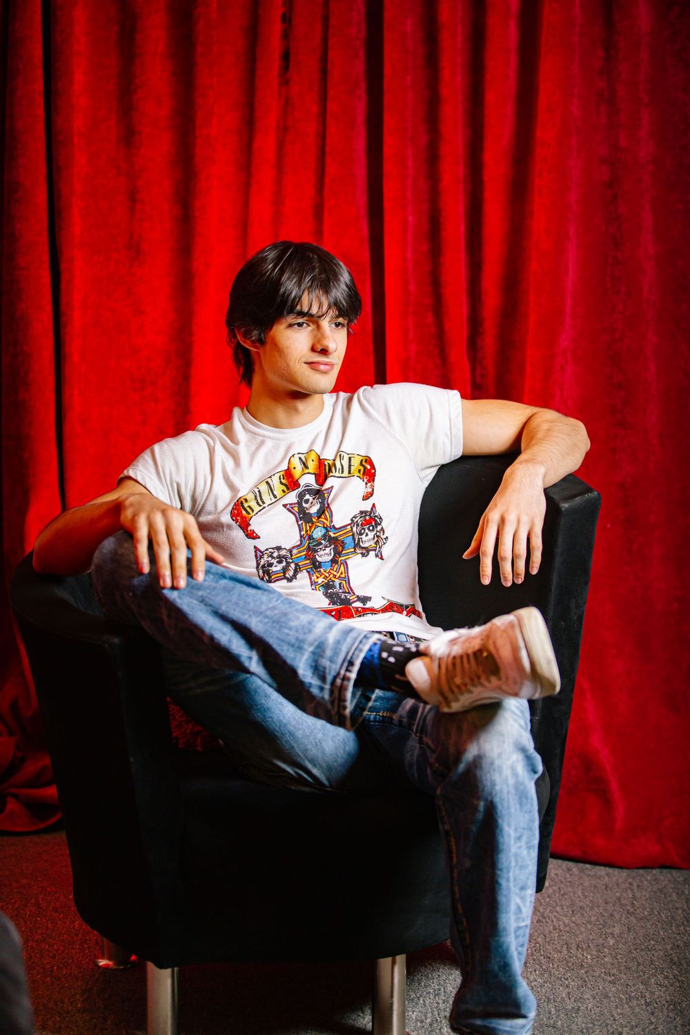 A person in a white graphic t-shirt and jeans sits casually in a black chair against a red curtain backdrop.