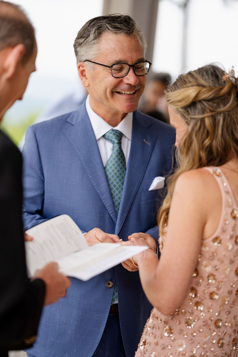 Smiling man in blue suit and green tie exchanges rings during an outdoor wedding ceremony.