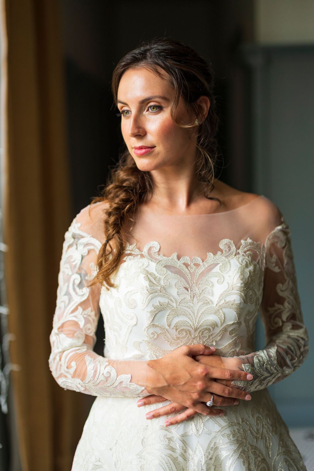 Elegant bride in long-sleeve lace off-shoulder wedding gown posing by window.