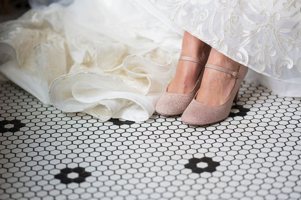 Bride wearing blush pink ankle strap heels beneath a ruffled white wedding gown on a classic black and white mosaic tile floor.