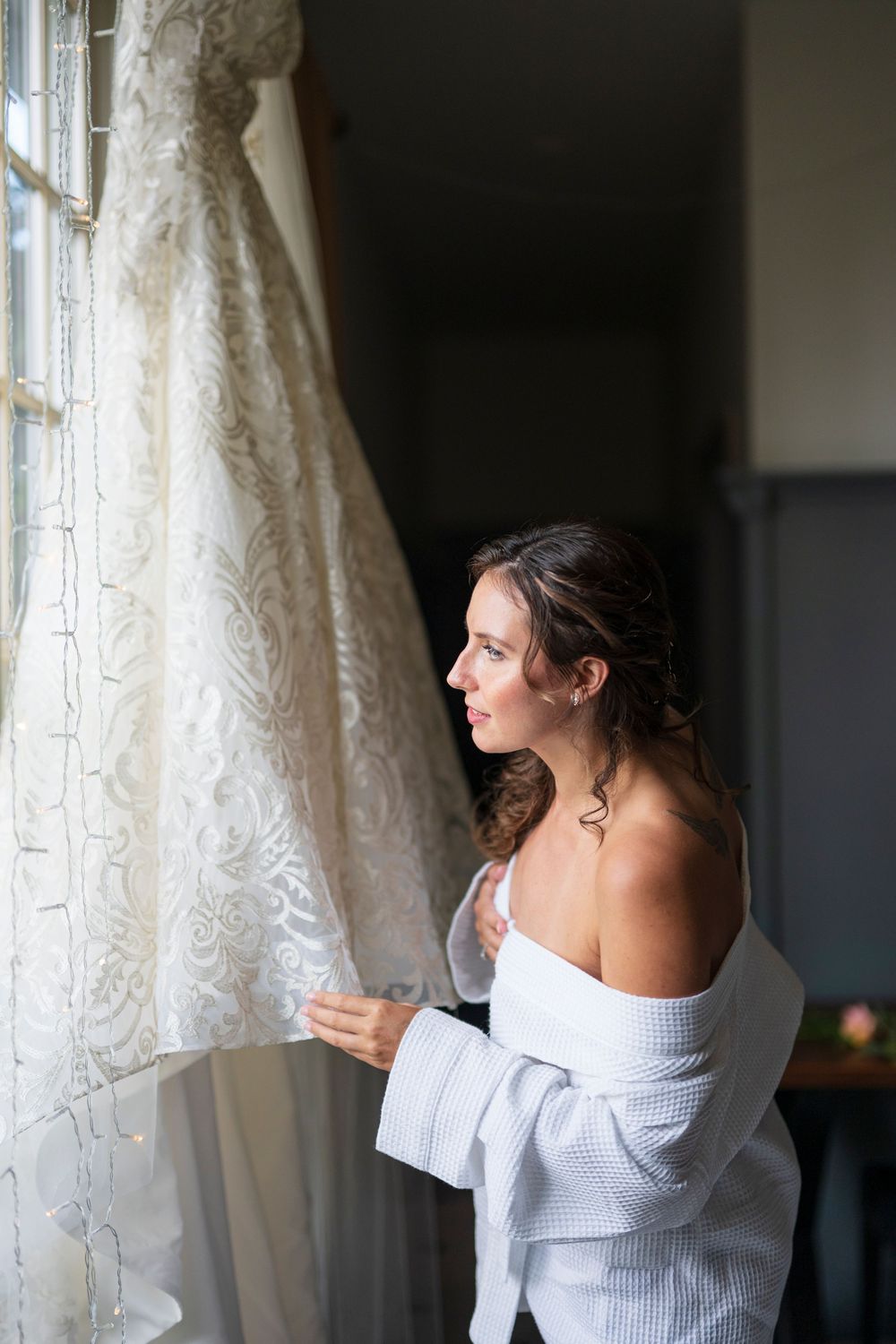 Bride in robe gazing at her lace wedding dress hanging by a window.