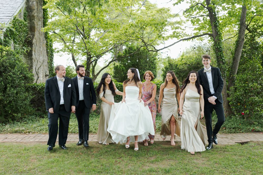 Wedding party walks together outdoors in lush garden setting, bride in white gown with bridesmaids in champagne dresses.