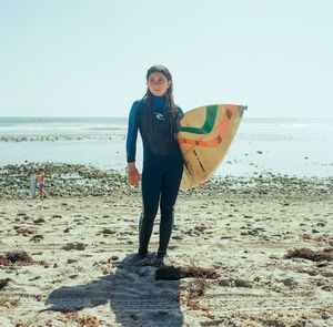 Young woman in black wetsuit holding colorful surfboard on sandy beach with calm ocean in background.